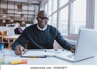 Portrait Of Confident Young Man At His Desk With Laptop Doing Paperwork. Happy African Man Looking At Camera While At Work.