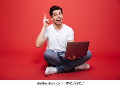 Portrait of a confident young man in white t-shirt holding laptop computer while sitting on a floor and pointing finger up isolated over red background