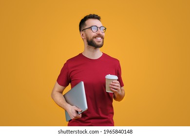 Portrait Of Confident Young Smiling Bearded Man In Casual Red T-shity And Trendy Spectacles Holding Laptop And Paper Cup Of Coffee, Isolated On Yellow Background.
