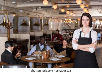 Portrait Of Confident Young Waitress With Arms Crossed In Cozy Busy Restaurant