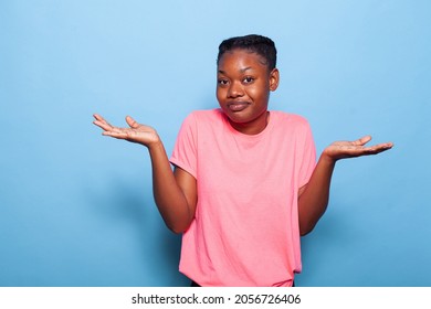 Portrait of confused african american teenager posing in pink t-shirt on a blue background in studio making uncertain facial expression. Young woman shrugging shoulders. I dont know concept - Shutterstock ID 2056726406