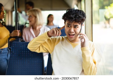 Portrait of confused asian guy talking on the cell phone and holding finger in ear while commuting by public transport, male can't hear because of noise and passengers, bad cellular connection