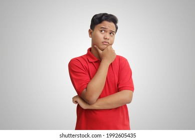 Portrait Of Confused Asian Man In Red Polo Shirt Standing Against Gray Background, Thinking About Question With Hand On Chin