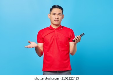 Portrait Of Confused Asian Young Man Using Mobile Phone And Looking At Camera Isolated On Blue Background