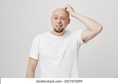 Portrait of confused bald caucasian male model with stupid expression, scratching head and looking aside not having any clue, standing over gray background. Man forgot something but do not know what