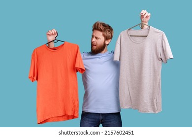 Portrait of confused bearded man with frowning face holding two hangers with gray and orange T-shirts, hard to choose the best attire. Indoor studio shot isolated on blue background.