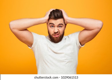 Portrait of a confused bearded man holding hands on his head isolated over yellow background
