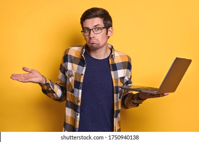 Portrait of confused frustrated man in glasses and casual clothes holding laptop computer on yellow studio wall, making don't know gesture, doesn't understand. Human emotion and technology concept