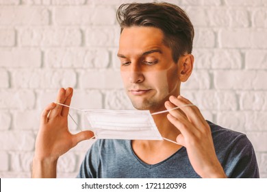 Portrait Of Confused Hipster Man With Funny Sunburn Tan Skin Wearing Medical Face Mask. Young Angry Man With Sun Tan Lines On Face From Protective Face Mask. UV Exposure. COVID-19 Pandemic Quarantine