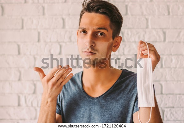 Portrait of confused hipster man with sunburn\
tanned face skin holding protective mask in hand. Young man patient\
with sun tanned lines on face after wearing medical face mask.\
COVID-19 quarantine