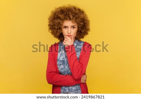 Portrait of confused lovely woman with Afro hairstyle holds chin, looks thoughtfully at camera, being deep in thoughts, thinking about work. Indoor studio shot isolated on yellow background.
