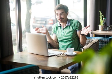 Portrait of confused man employee wearing green T-shirt, working online on laptop, shrugging shoulders, making no idea gesture, whatever. Indoor shot near big window, cafe background. - Powered by Shutterstock