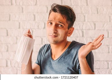 Portrait Of Confused Man With Funny Sun Tan Lines On Face Holding Protective Mask In Hand. Young Hipster Man With Tanned Sunburn Skin After Wearing Medical Face Mask. COVID-19 Coronavirus Prevention