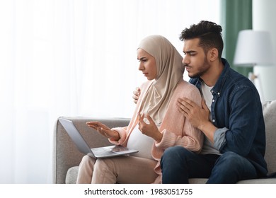 Portrait Of Confused Pregnant Muslim Couple Looking At Laptop Screen At Home, Frustrated Islamic Spouses Awaiting For Baby Using Computer While Sitting Together On Couch In Living Room, Copy Space