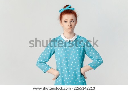 Portrait of confused puzzled ginger woman standing with hands on hips, looking away, thinking, keeps hands on hips, wearing blue dress. Indoor studio shot isolated on gray background.