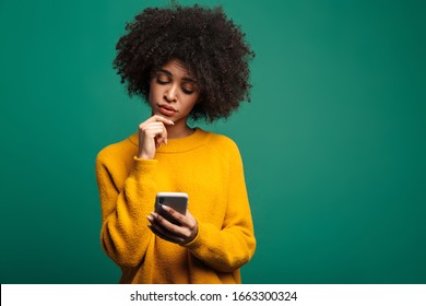 Portrait of a confused young african woman wearing sweater standing isolated over green background, using mobile phone - Powered by Shutterstock