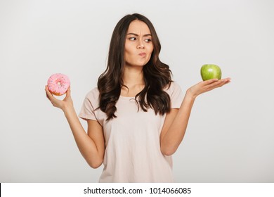 Portrait of a confused young asian woman choosing between donut and green apple isolated over white background