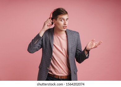 Portrait of a confused young attractive man wearing jacket standing isolated over pink background, lstening to loud music with wireless headphones