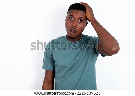 Portrait of confused young handsome man wearing green T-shirt over white background holding hand on hair and frowning, panicking, losing memory. Worried and anxious can not remember anything.