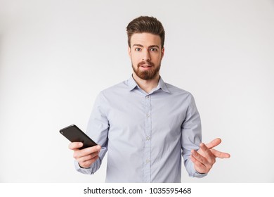 Portrait Of A Confused Young Man Dressed In Shirt Holding Mobile Phone Isolated Over White Background