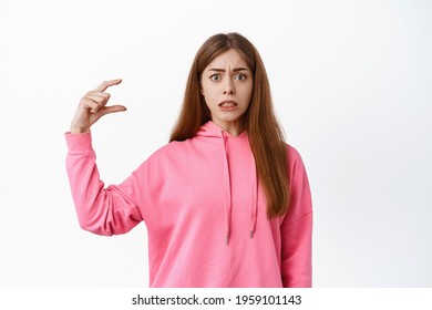 Portrait Of Confused Young Woman Showing Tiny Little Thing Gesture, Looking Nervous At Camera, Display Small Object With Anxious Face, Standing Over White Background.