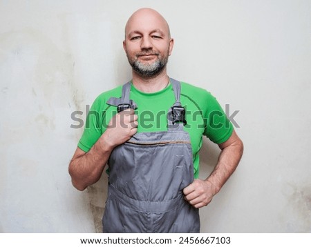 Portrait of a construction worker on light wall background. Builder in grey heavy duty clothes and green shirt. Male in 40s, bald with grey beard. Lean athletic type. Friendly face