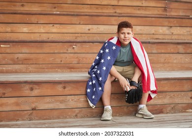 Portrait Of Content College Black Baseball Player Wrapped Into American Flag Sitting On Wooden Bleacher Bench