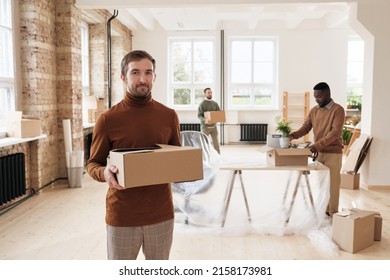 Portrait Of Content Handsome Young Manager In Turtleneck Holding Closed Stuff Box Against Colleagues Setting Up New Office