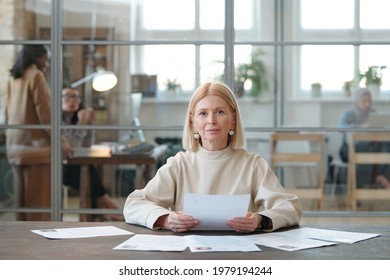 Portrait of content mature HR manager with blond hair sitting at table and reading CVs of candidates in modern open space office