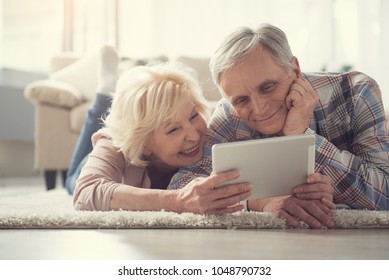 Portrait of content pensioners relaxing on soft rug in living room and looking at tablet - Powered by Shutterstock