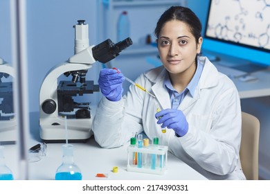 Portrait of content young Indian female pharmacist in lab coat and surgical gloves sitting at desk and mixing reagents in test tube using pipette - Shutterstock ID 2174390331