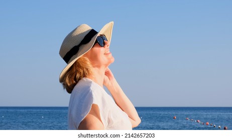 Portrait of a contented 50 year old woman wearing a straw hat and sunglasses enjoying the sun against the blue sea. Summer, vacation, vacation, active retirees - Powered by Shutterstock