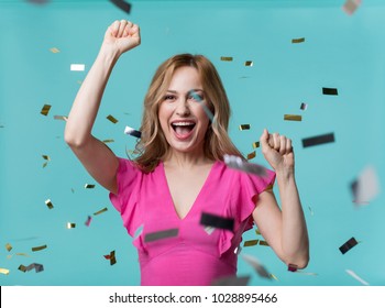 Portrait of contented blonde girl exulting while raising her hands up as gesture of triumph. Confetti flying in air. Isolated on blue background - Shutterstock ID 1028895466