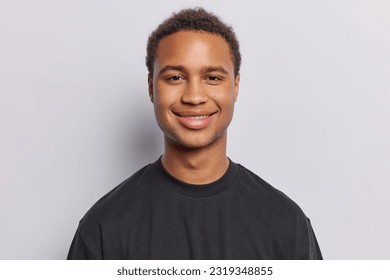 Portrait of contented dark skinned man with short curly hair radiates satisfaction upon hearing good news exudes pure satisfaction wears black t shirt isolated over white background. Emotions concept