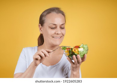 Portrait of a contented mature woman in a white shirt holding a plate of salad and a fork, ready for dinner. Studio shot on a yellow background