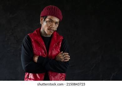 Portrait Of Cool Asian Hip Hop Young Bearded Man With Tattoo On Face, Neck And Hands Standing And Cross His Arms With Nose Piercing, Big Ear Piercing And Wear Red Knit Hat And Vest. Black Background