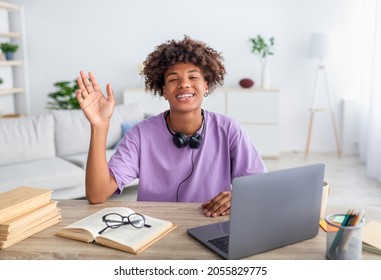 Portrait Of Cool Black Teen Guy With Headphones, Laptop And Study Materials Waving At Camera, Sitting At Desk Indoors. Funky Black Teenager Learning Online At Home. Web Based Education Concept