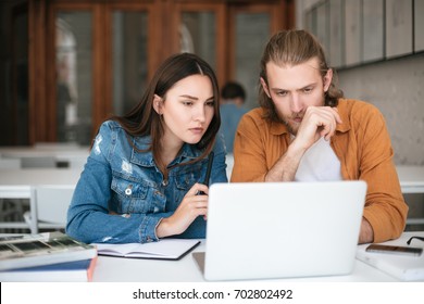 Portrait Of Cool Boy And Girl Thoughtfully Looking On Laptop. Young Students Sitting In Classroom And Studying Together With Laptop And Books On Table. Man And Woman Working On Laptop In Office