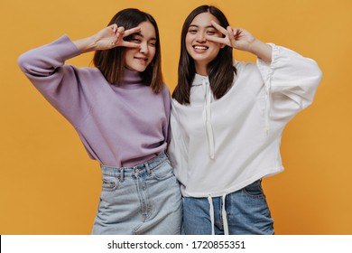 Portrait Of Cool Brunette Asian Girl Shows V-signs On Isolated. Happy Woman In Jeans And White Hoodie Poses On Orange Background. Pretty Lady In Denim Skirt And Purple Sweater Smiles.