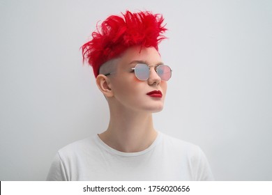 Portrait Of A Cool Fashionable Modern Young Girl. A Short Haircut With Shaved Temple. Dyed Bright Red Hair. Red Lipstick. Studio Photo On A White Background. Suspenders On Checkered Pants And