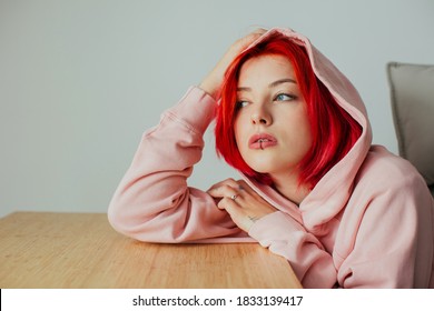 Portrait of a cool female with bright red hair, lip piercing and hoodie holding head on desk looking out the window - Shutterstock ID 1833139417