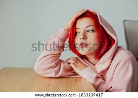 Portrait of a cool female with bright red hair, lip piercing and hoodie holding head on desk looking out the window