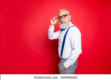 Portrait Of Cool, Manly, Fashionable Old Man Holding Hand In Pocket And Eyelet Of Glasses On His Face, Looking At Camera With Smirk, Isolated On Red Background, Having Blue Suspenders, Pants