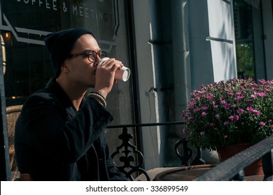 Portrait Of Cool Stylish Handsome Lonely Hipster Guy Wearing All Black In Hat And Glasses Sitting In Cafe Drinking Coffee During Sunny Morning