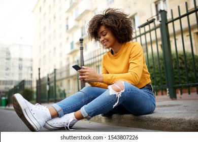 Portrait of cool young african american woman sitting outside on street with cellphone