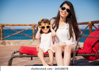 Portrait Of A Cool Young Mom And Her Little Girl Wearing Sunglasses And Relaxing On The Beach On A Sunny Day