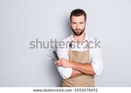 Portrait with copy space, empty place of virile harsh barber having his arms crossed, holding equipments in hand, looking at camera, isolated on grey background