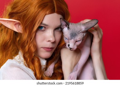 Portrait Of Cosplay Elf Young Redhead Woman With Sphynx Kitten On Red Background. Elf In White Dress, With Gorgeous Wavy Hair, Leaned Her Head Against Kitten's Head, Looks At Camera With Stern Gaze.