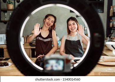 Portrait Of Couple Girl Baker Smiling And Waving Hand At Camera On Portable Ring Light Tripod In The Kitchen.