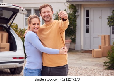 Portrait Of Couple Holding Keys Outside New Home On Moving Day Unloading Boxes From Car